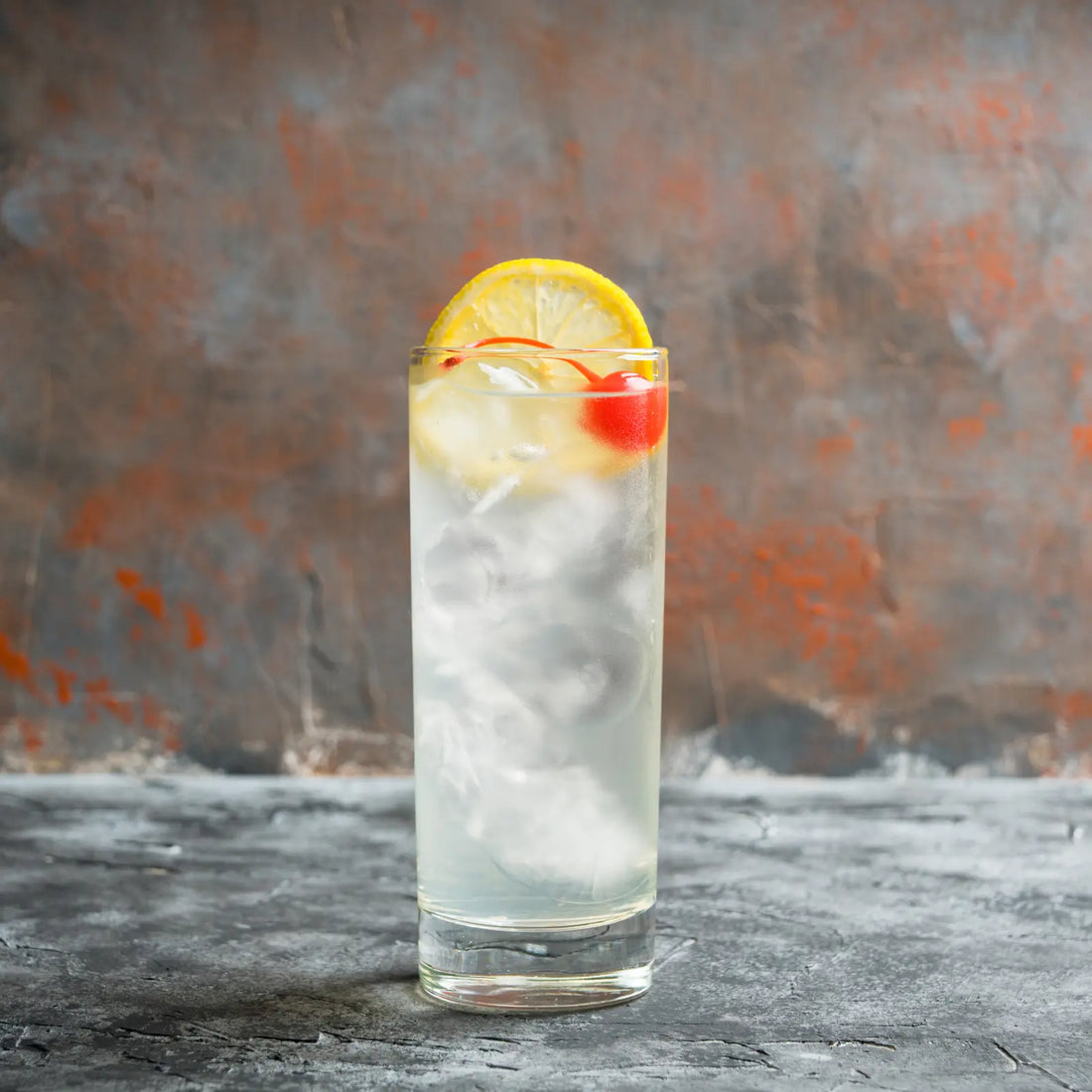 Refreshing Tom Collins cocktail with a cherry and lemon garnish, served against a textured orange-gray backdrop