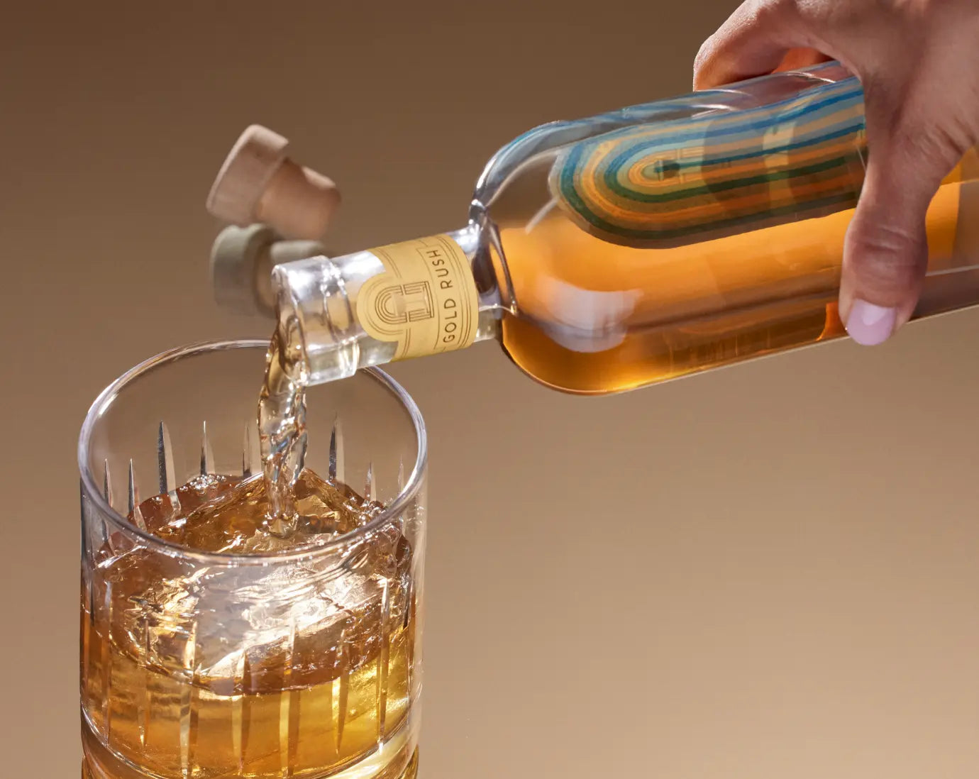 Premade cocktail being poured from a Gold Rush bottle into a glass filled with ice on a warm-toned background.