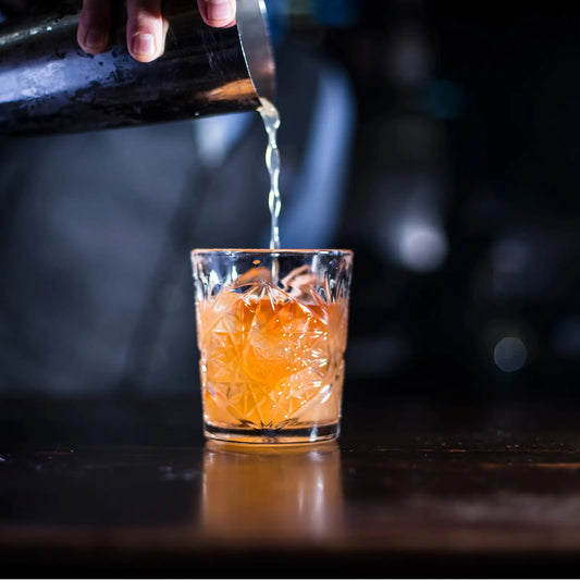 Bourbon Old Fashioned being poured from a shaker into an intricately designed glass with ice