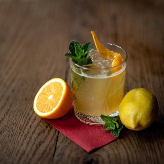 Bourbon smash cocktail garnished with orange peel, mint leaves, and served with fresh orange and lemon on a wooden table