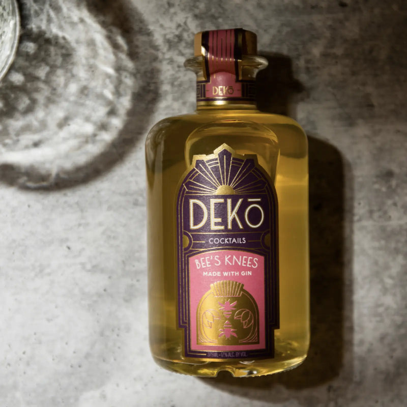 DEKO Bee's Knees gin cocktail bottle resting on a textured concrete surface.