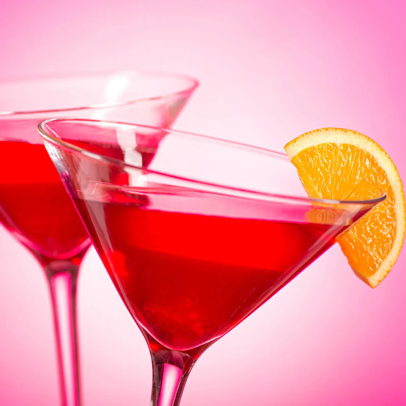 Two Cosmopolitan cocktails in martini glasses garnished with orange slices against a pink background.