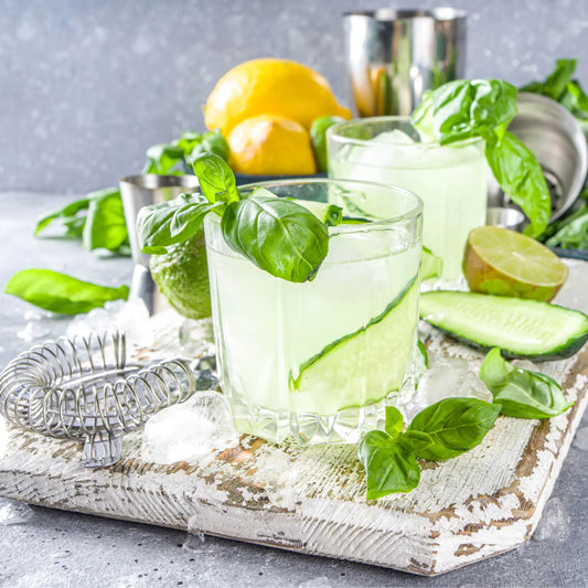 Vodka cucumber cocktails garnished with fresh basil leaves, lime, and ice on a rustic white serving board