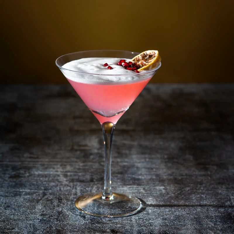 French Martini with frothy top, pomegranate seeds, and dried citrus in a stemmed glass.