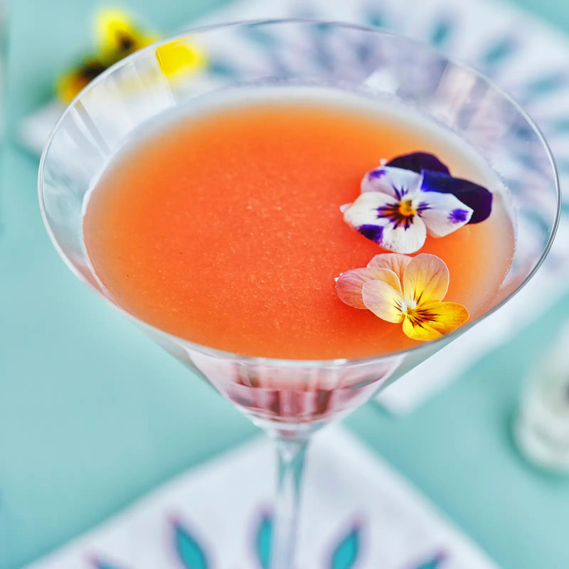 Pink grapefruit Greyhound cocktail in a martini glass garnished with edible pansy flowers.