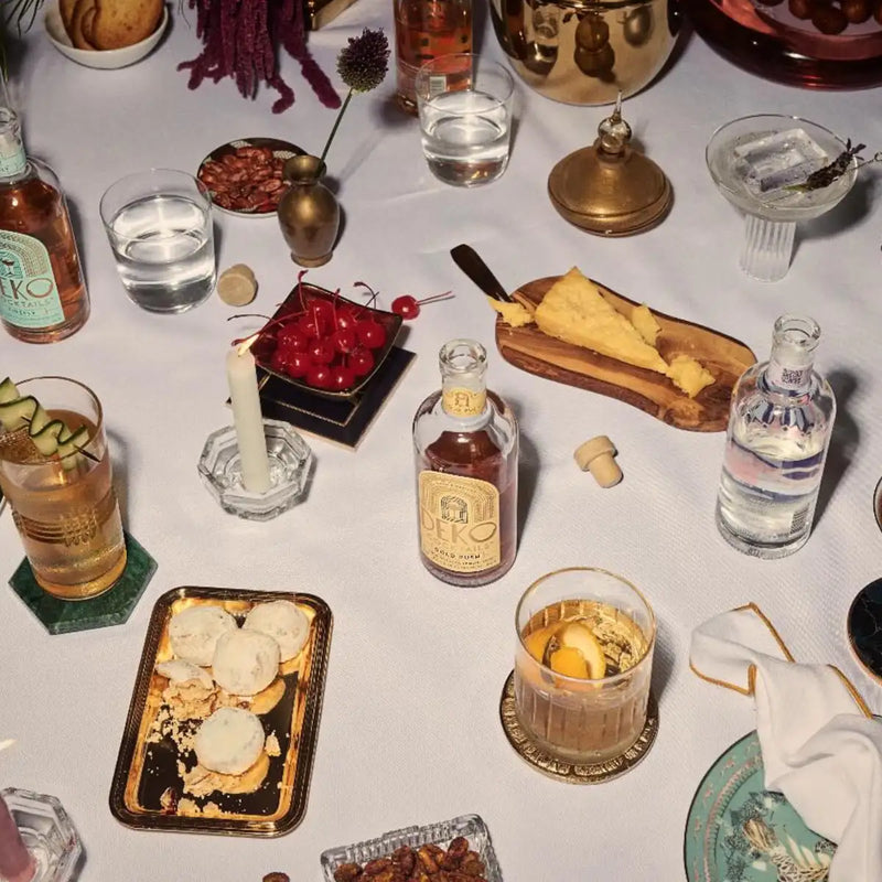 Ready-to-drink bottled cocktails and glasses spread across a white entertaining table with cheese and snacks.