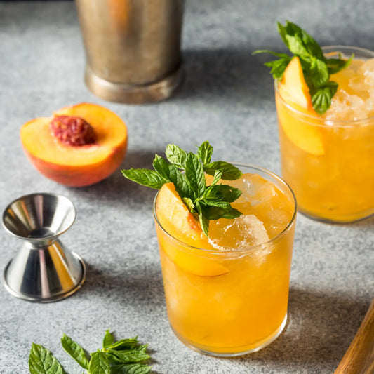 Peach bourbon cocktails with fresh mint, crushed ice, and a halved peach next to cocktail tools on a gray surface