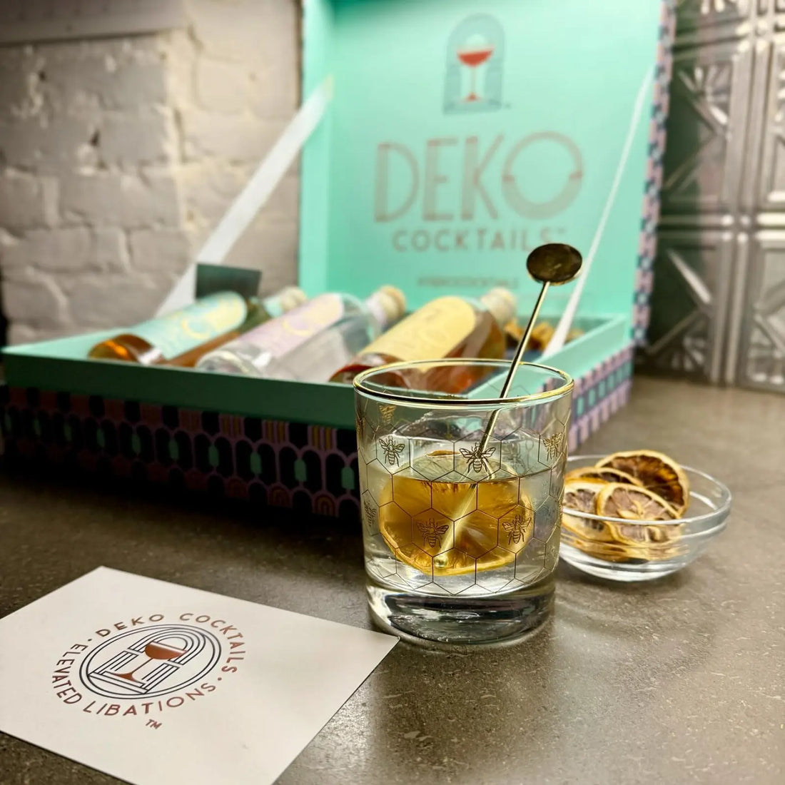 A glass of Deko Cocktails Bee's Knees garnished with dried citrus, displayed alongside a premium cocktail gift box.