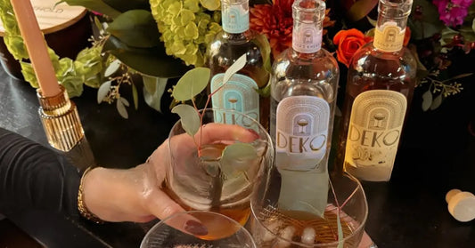 Bottled cocktails lineup of Deko surrounded by flowers and glasses in a celebratory toast.