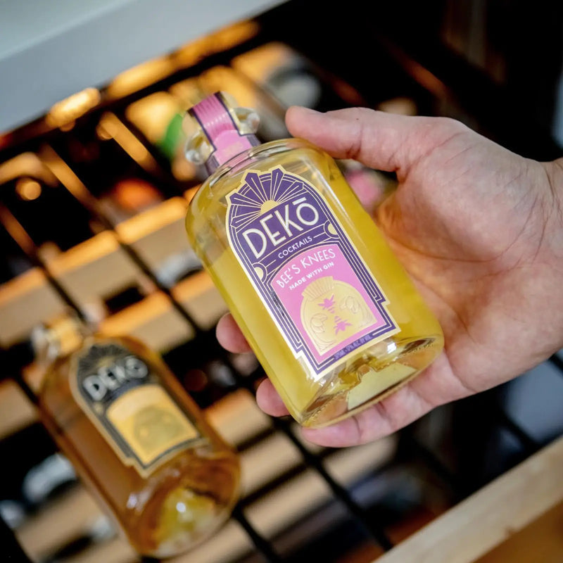 Hand holding a Deko Bee's Knees gin cocktail bottle near a wine rack.
