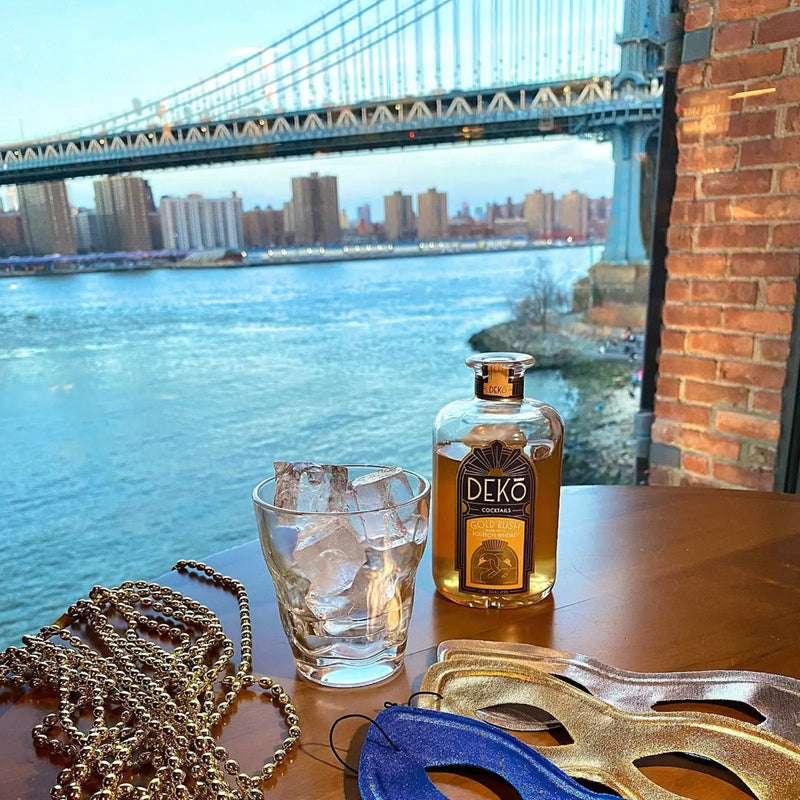 Deko Gold Rush bourbon cocktail bottle beside an iced glass and party masks by the Manhattan Bridge.