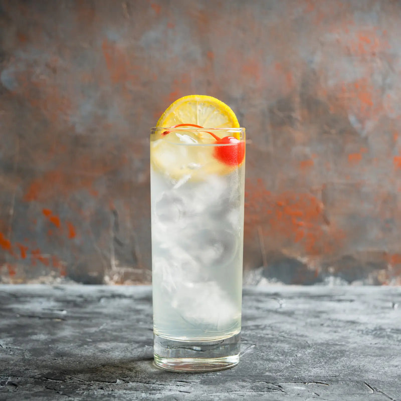 Refreshing Tom Collins cocktail with a cherry and lemon garnish, served against a textured orange-gray backdrop