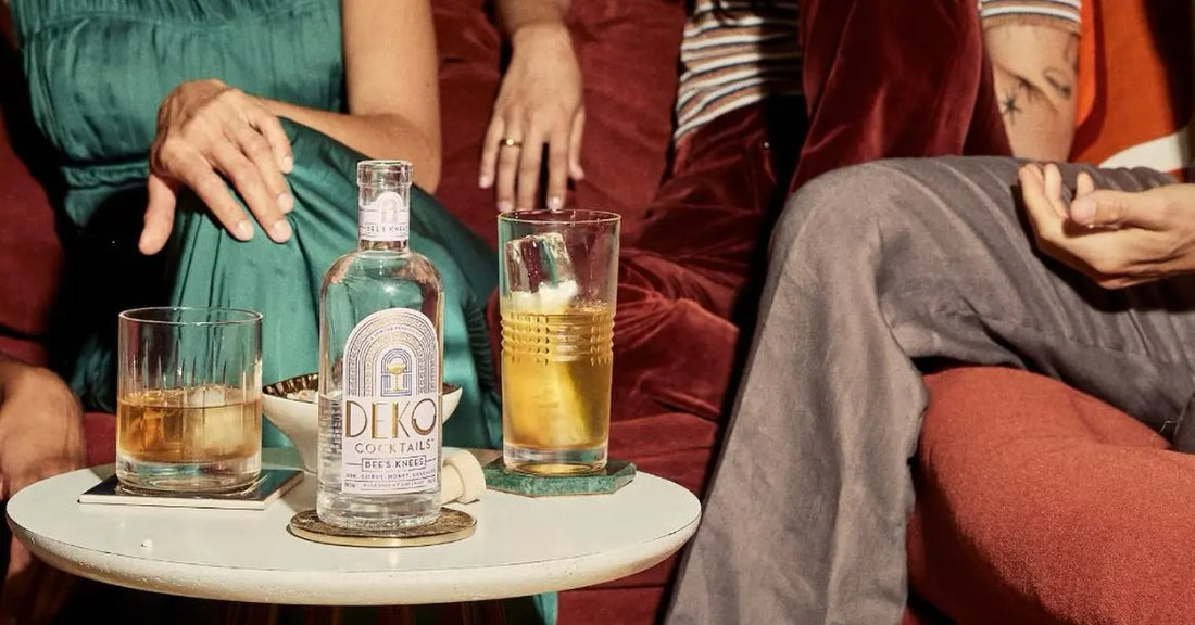 Bee's Knees gin cocktail bottle on a small table with mixed drinks, surrounded by elegantly dressed people on a velvet couch