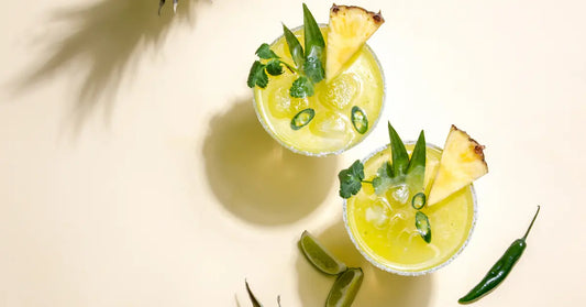 Jalapeno margarita in two glasses, garnished with pineapple, cilantro, and jalapeño slices, surrounded by fresh limes and chili peppers