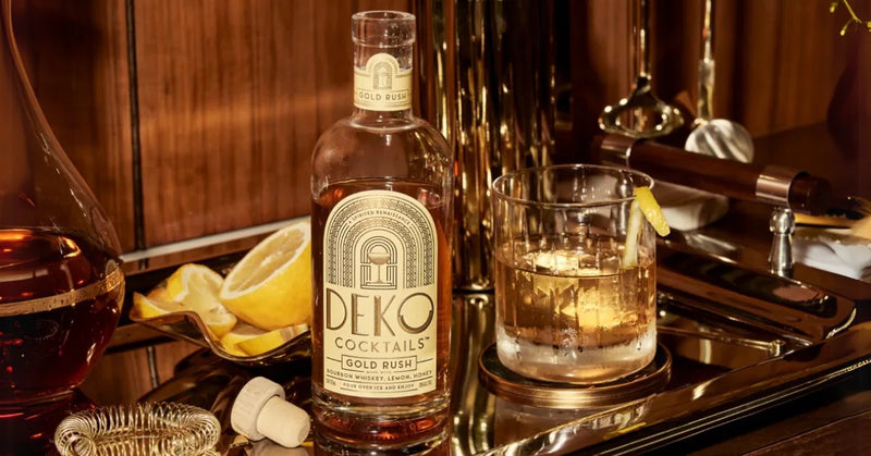 Deko Gold Rush bourbon cocktail poured over ice beside its bottle, lemon, and bar tools.