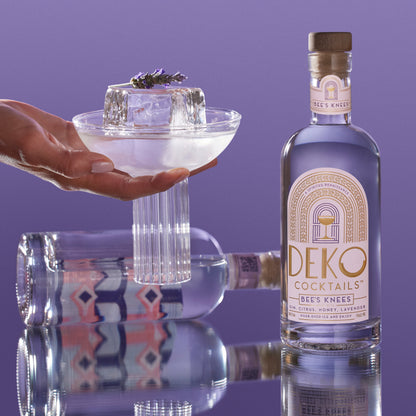 Bee’s Knees cocktail featuring a bottle of Deko Cocktails Bee’s Knees made with gin, citrus, honey, and lavender, shown with a lavender-garnished glass on a reflective surface.