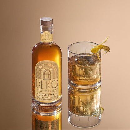 Gold Rush cocktail featuring a bottle of Deko Cocktails Gold Rush made with bourbon whiskey, lemon, and honey, shown next to a garnished glass over ice on a mirrored surface.