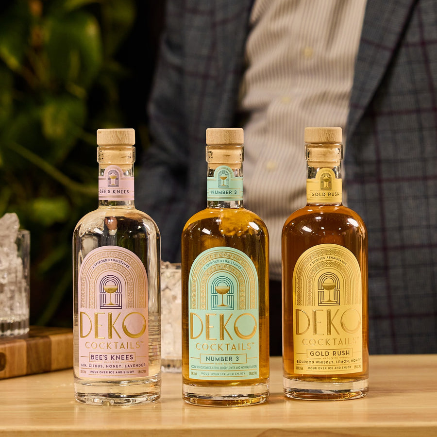 Deko Cocktails 3 Pack with Bee's Knees, Number 3 and Gold Rush bottles on a bar top beside a glass of ice.