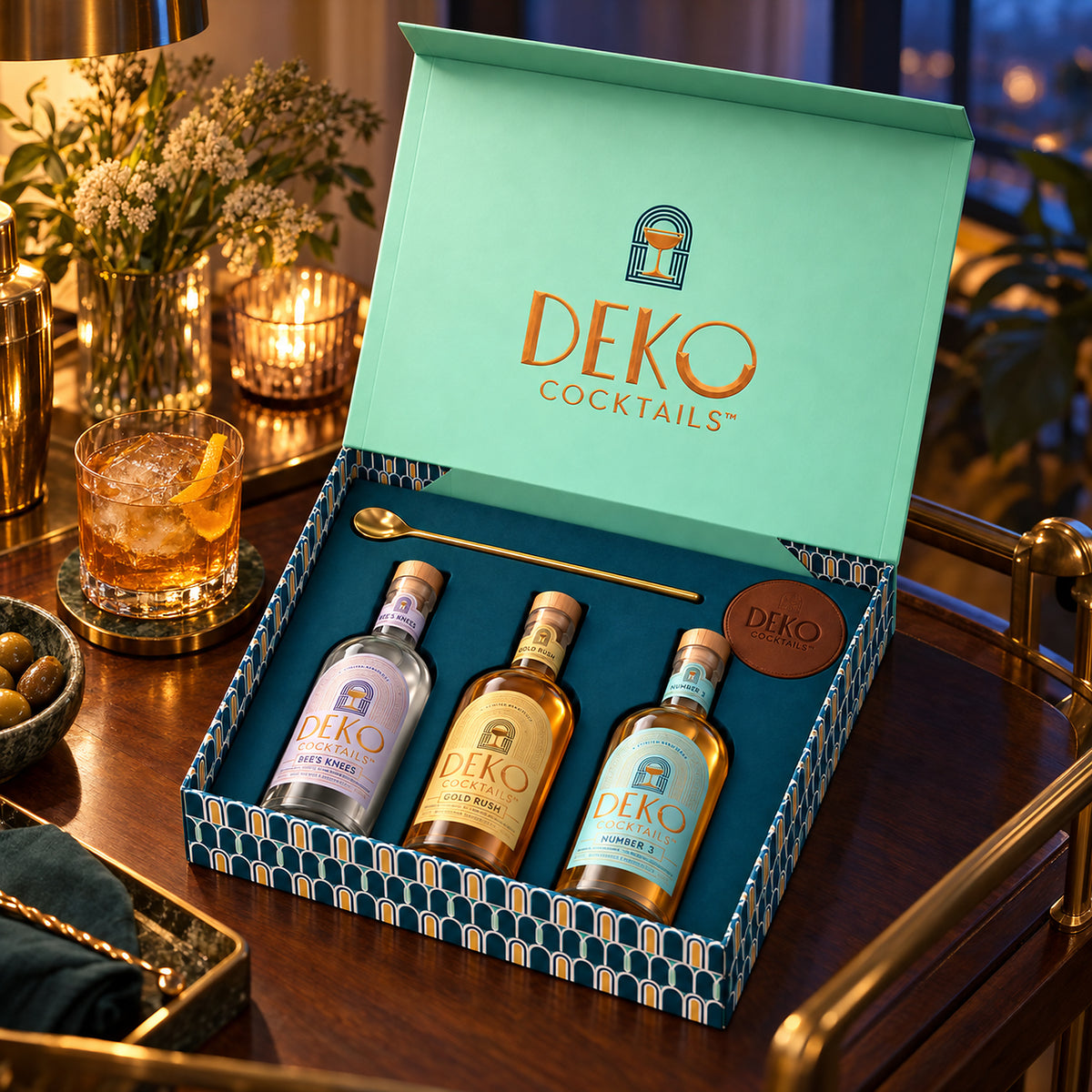 Deko Cocktails gift box open on a bar cart, showing Bee’s Knees, Gold Rush and Number 3 with a gold bar spoon and vegan leather coaster
